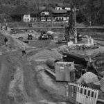 Monochrome image of construction site next to a river with heavy machinery and workers.
